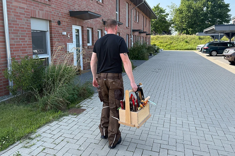 kosten-montageservice Ein Mann in braunen Arbeitshosen und einem schwarzen Shirt trägt einen Werkzeugkasten auf dem Gehweg neben einem Backsteingebäude zu einem Eingang.